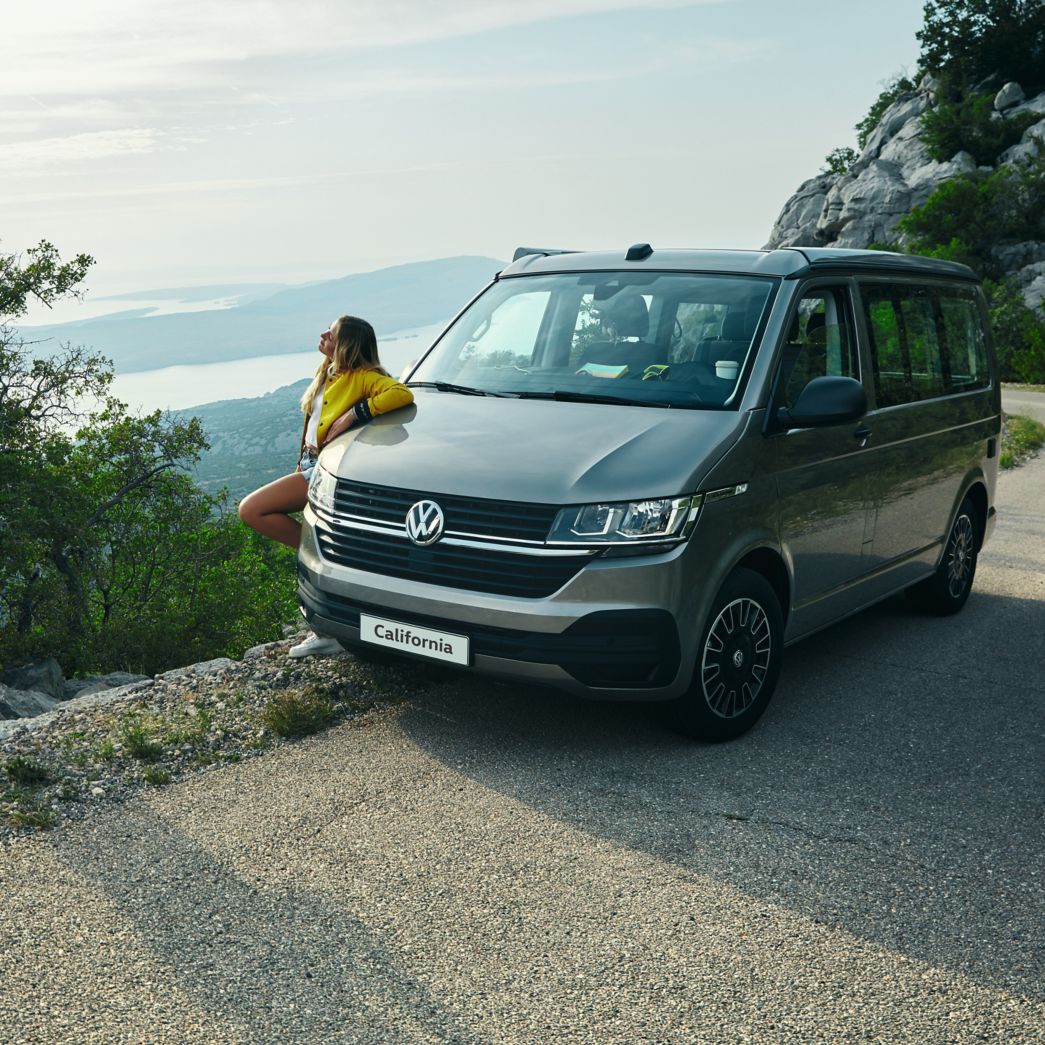VW California Beach an der Seite einer Bergstrasse mit Frau daneben