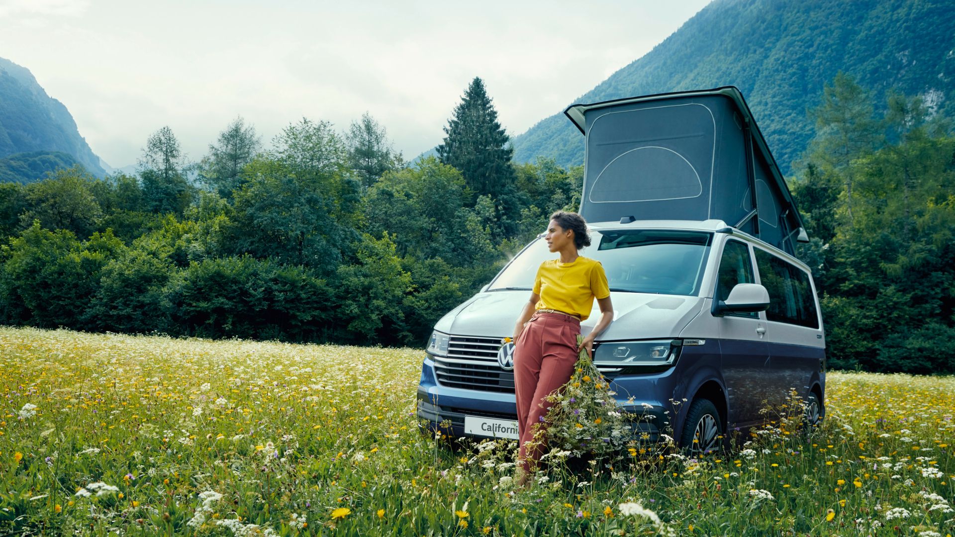 VW California Ocean front view on a meadow with flowers with woman leaning against it