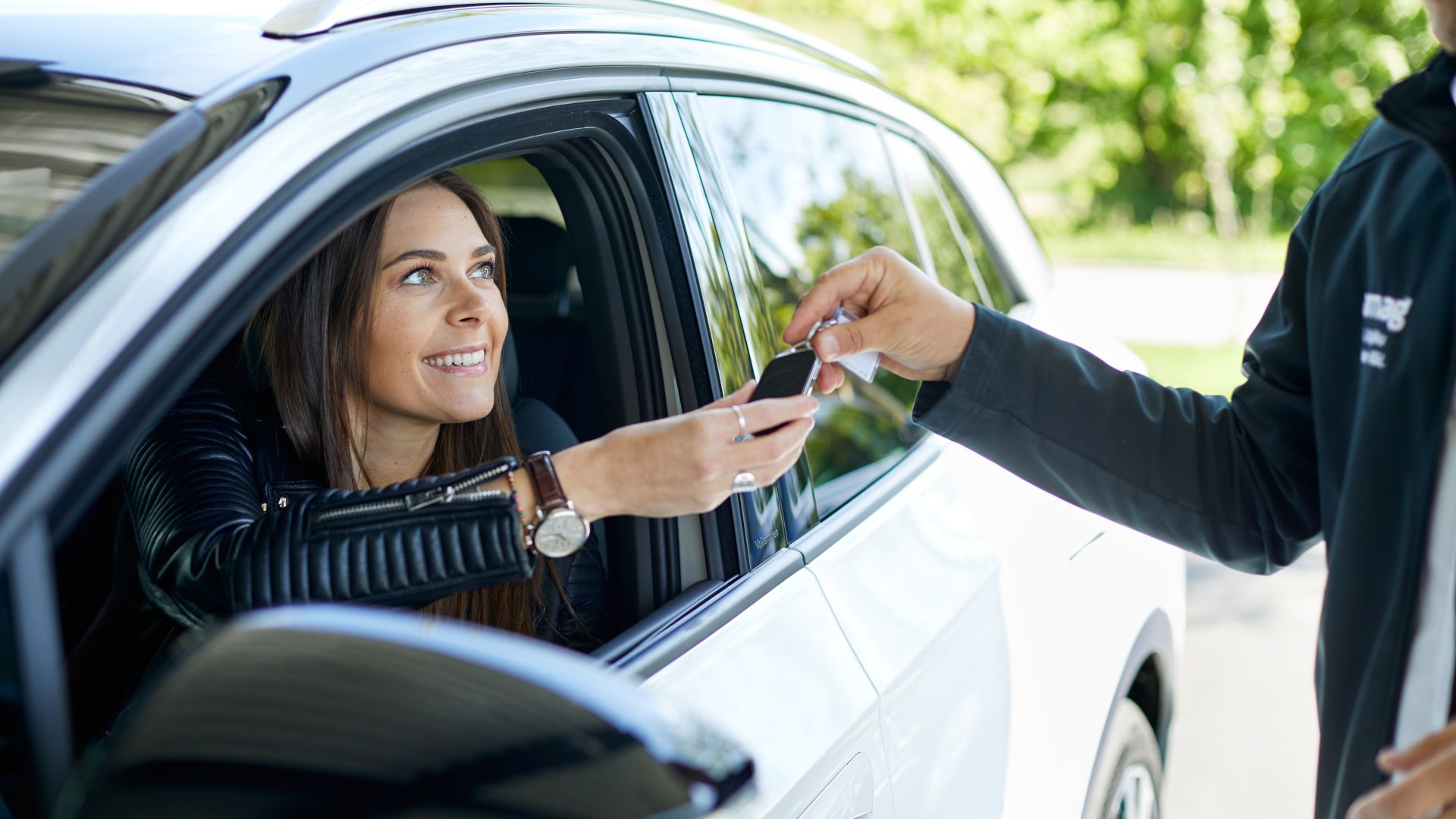Une femme souriante dans une voiture blanche reçoit les clés de voiture d'une personne qui passe par la fenêtre ouverte.