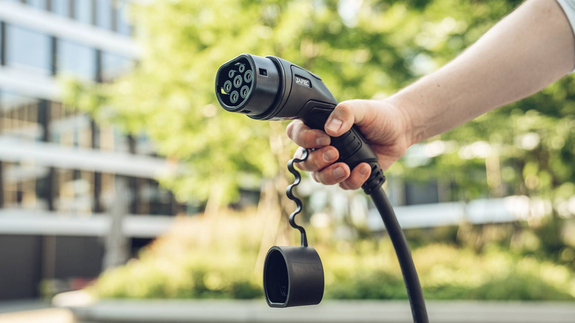 Une main tient une prise de charge noire pour voitures électriques, avec un arrière-plan flou d'arbres et de bâtiments.