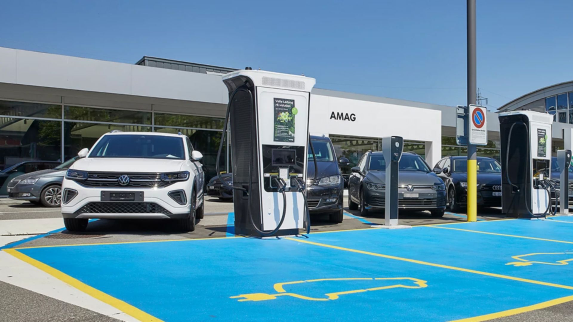 Des voitures électriques sont garées sur des places bleues, près de bornes de recharge rapide blanches, devant un bâtiment AMAG.