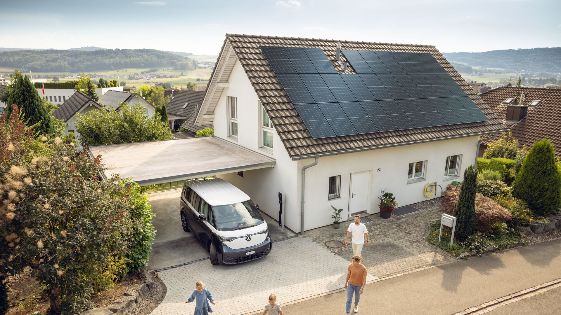 Une maison moderne avec des panneaux solaires sur le toit, devant une fourgonnette électrique sous un carport et une famille dans le jardin de devant.