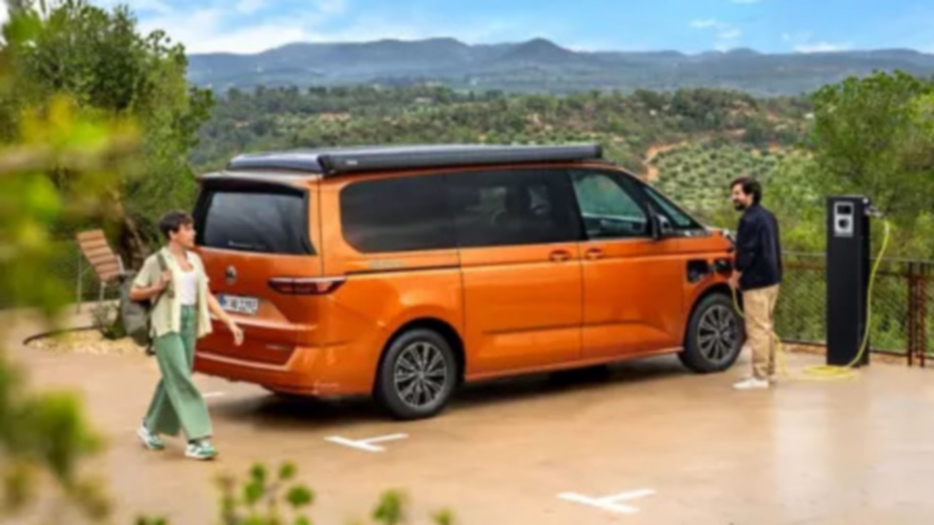 Vista laterale e posteriore del VW California eHybrid arancione in un parcheggio durante il caricamento